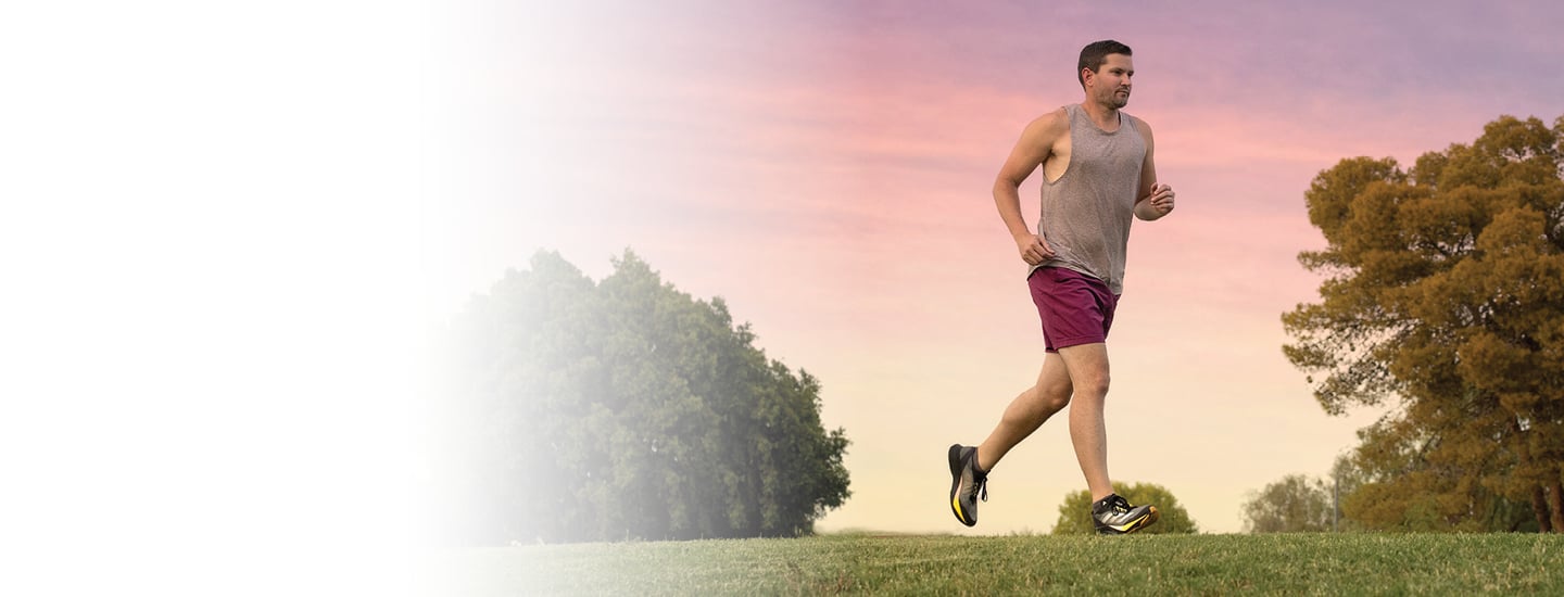 A man wearing a gray tank top and maroon running shorts runs across a grass field with trees in the background.